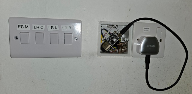 Left to right, low down on the wall: Double gang fitting with 4 light switches labelled (FB M, LR C, LR L, LR R). Single gang box with ESP32-C6 inside behind a transparent plastic cover. Single gang socket with power supply.