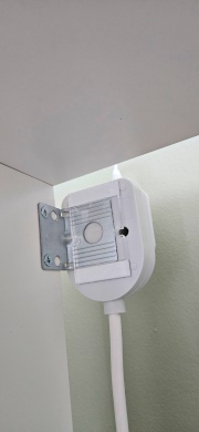 [Upside-down white power extension socket attached to metal IKEA KALLAX wall bracket using pieces of acrylic superglued to the back to make a slightly raised holding plate, with a white cable running down and plug hidden from view inserted into it]