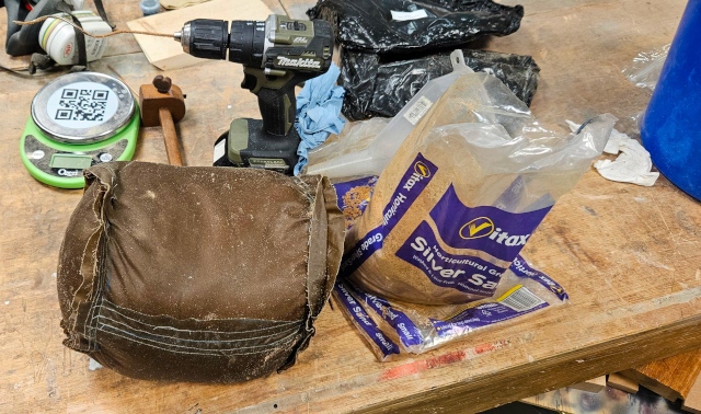 [Brown waxed canvas sewn in a cylinder shape containing ~11kg of sand, with a drill behind it containing a twisted piece of welding rod, next to an almost empty bag of sand on top of other empty bags of sand]
