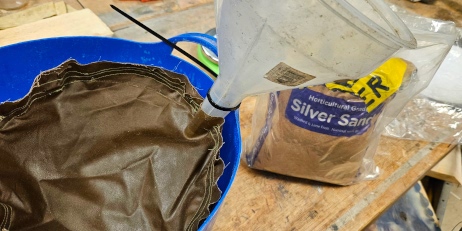[Brown waxed canvas sewn in a cylinder shape, with a funnel inside it, inside a blue bucket, next to a full 5kg bag of sand]