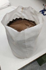 [Brown waxed canvas doorstop sitting inside white ripstop PVC sewn in a cylinder shape (as a cover) that's open at the top]